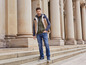 Un homme avec un gilet, un sweat à capuche et un jean pose devant un bâtiment avec des colonnes.