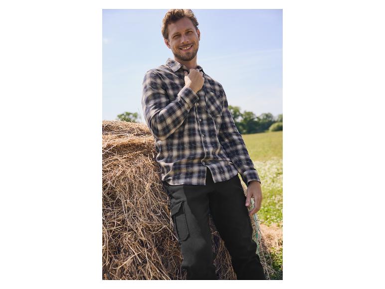 Homme en chemise à carreaux et pantalon noir posant près de bottes de foin.