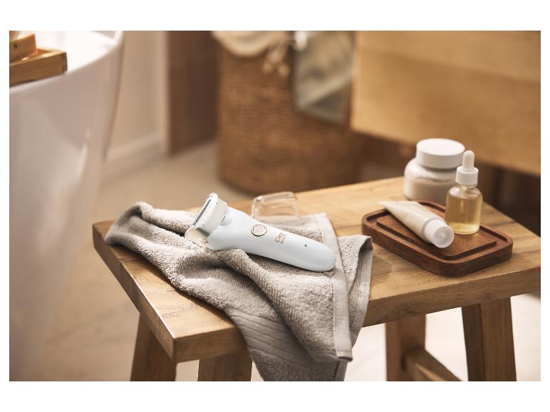 Épilateur pour femme sur une serviette, à côté de produits de soin sur un tabouret en bois.