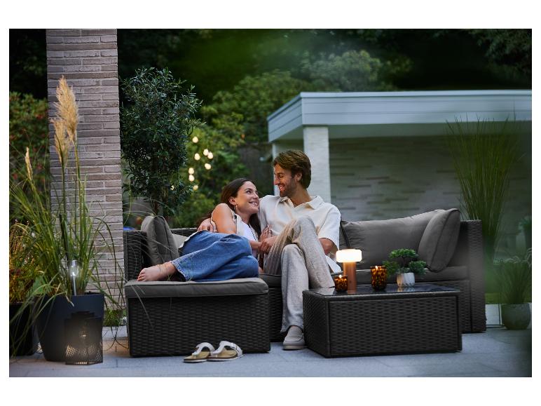 Couple se détendant sur un canapé d'extérieur avec coussins, entouré de plantes et d'un éclairage chaleureux.