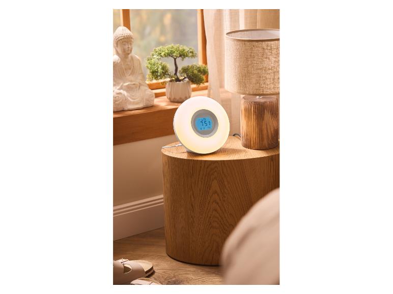 Réveil lumineux sur table de chevet avec lampe et décorations, fenêtre en arrière-plan.