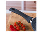 Une poêle noire avec une poignée en silicone sur une planche à découper en bois avec des tomates et un poivron rouge.