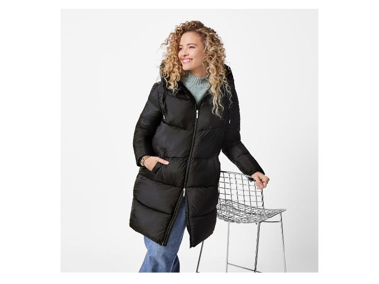 Femme souriante en longue doudoune noire, la main sur une chaise métallique.