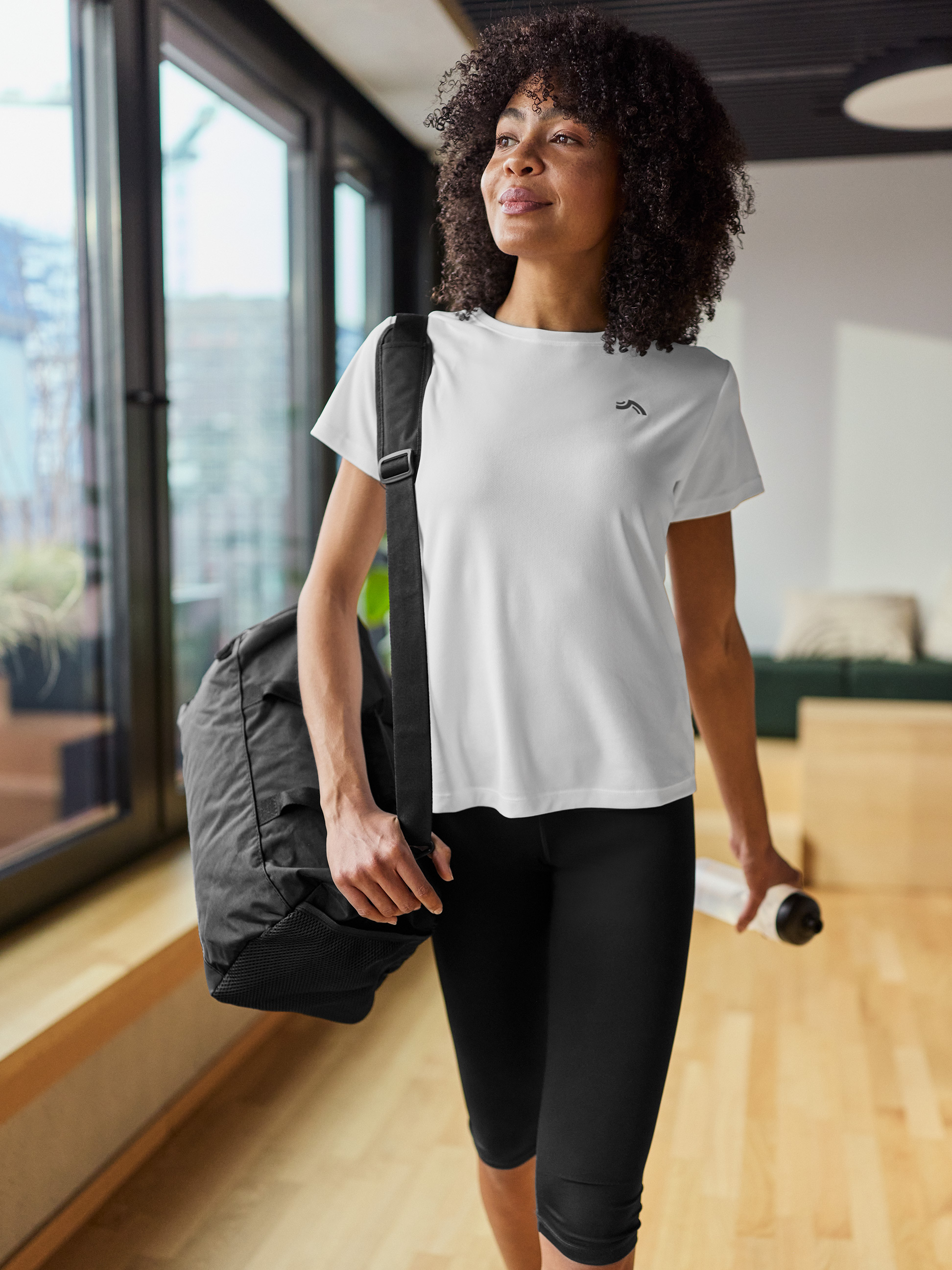 Femme en t-shirt de sport blanc, legging noir, avec sac de sport et bouteille d'eau.