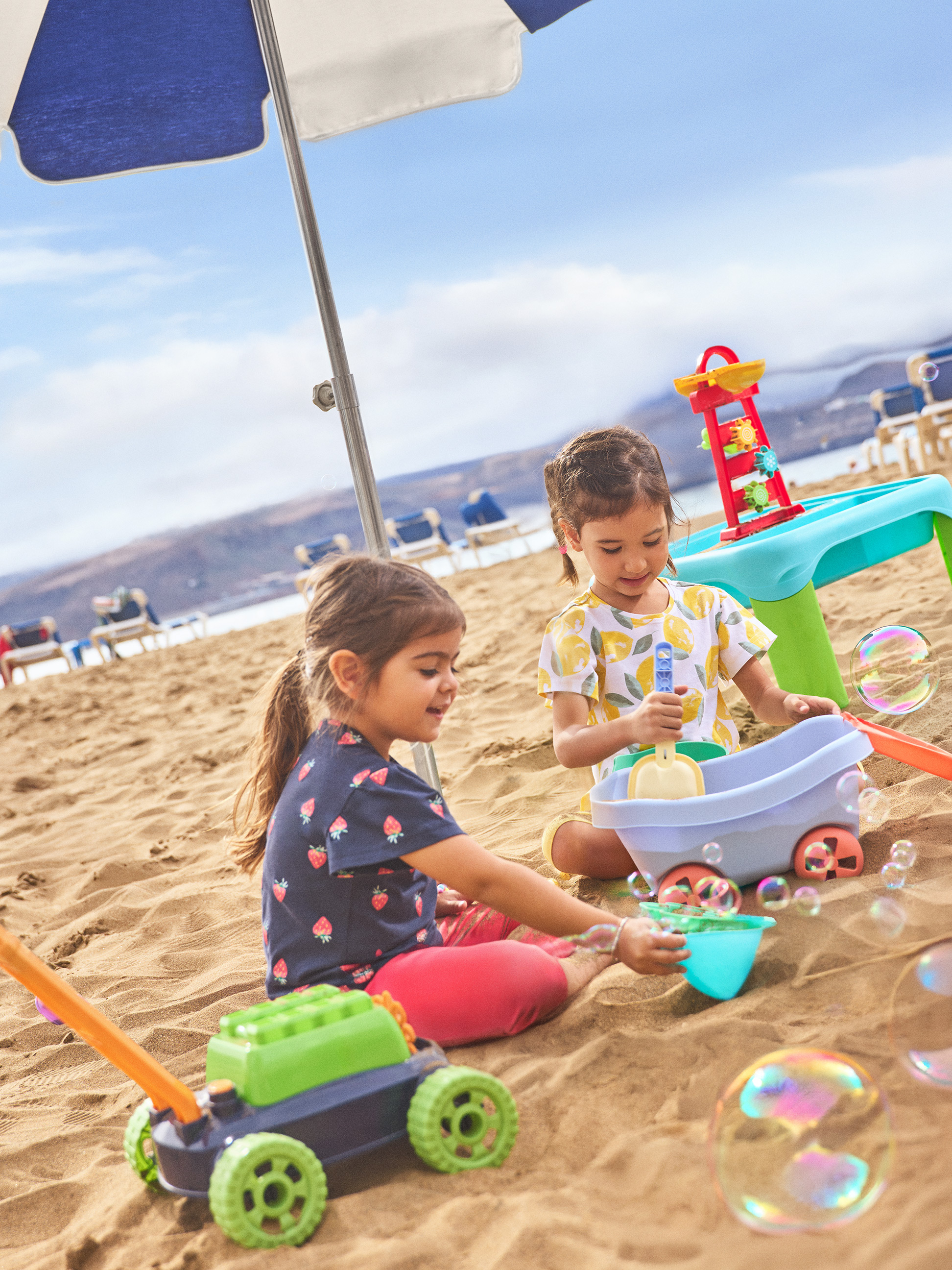 Deux petites filles jouent avec des jouets de plage et des bulles de savon sous un parasol.