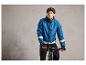 Homme en veste de vélo bleue avec des bandes réfléchissantes, un casque et des gants noirs sur un vélo noir.