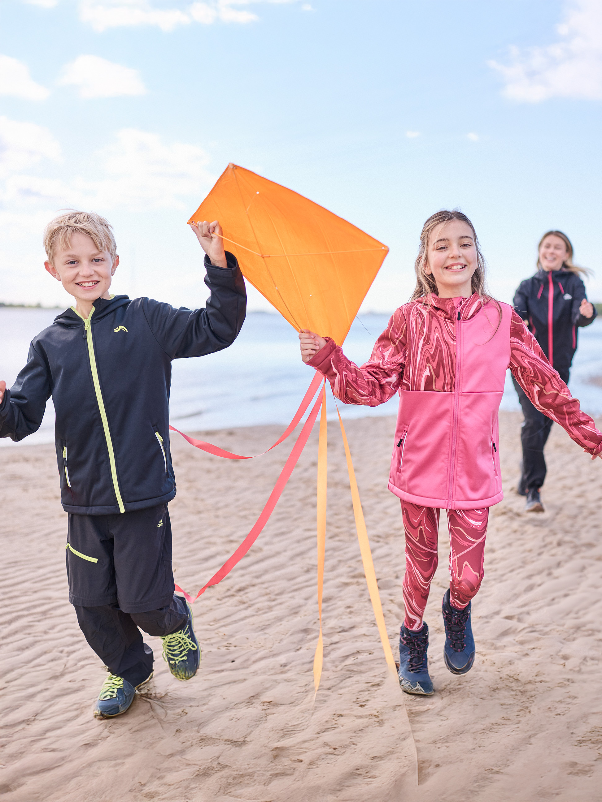 Enfants en tenue de sport courant sur une plage avec un cerf-volant, femme en arrière-plan.