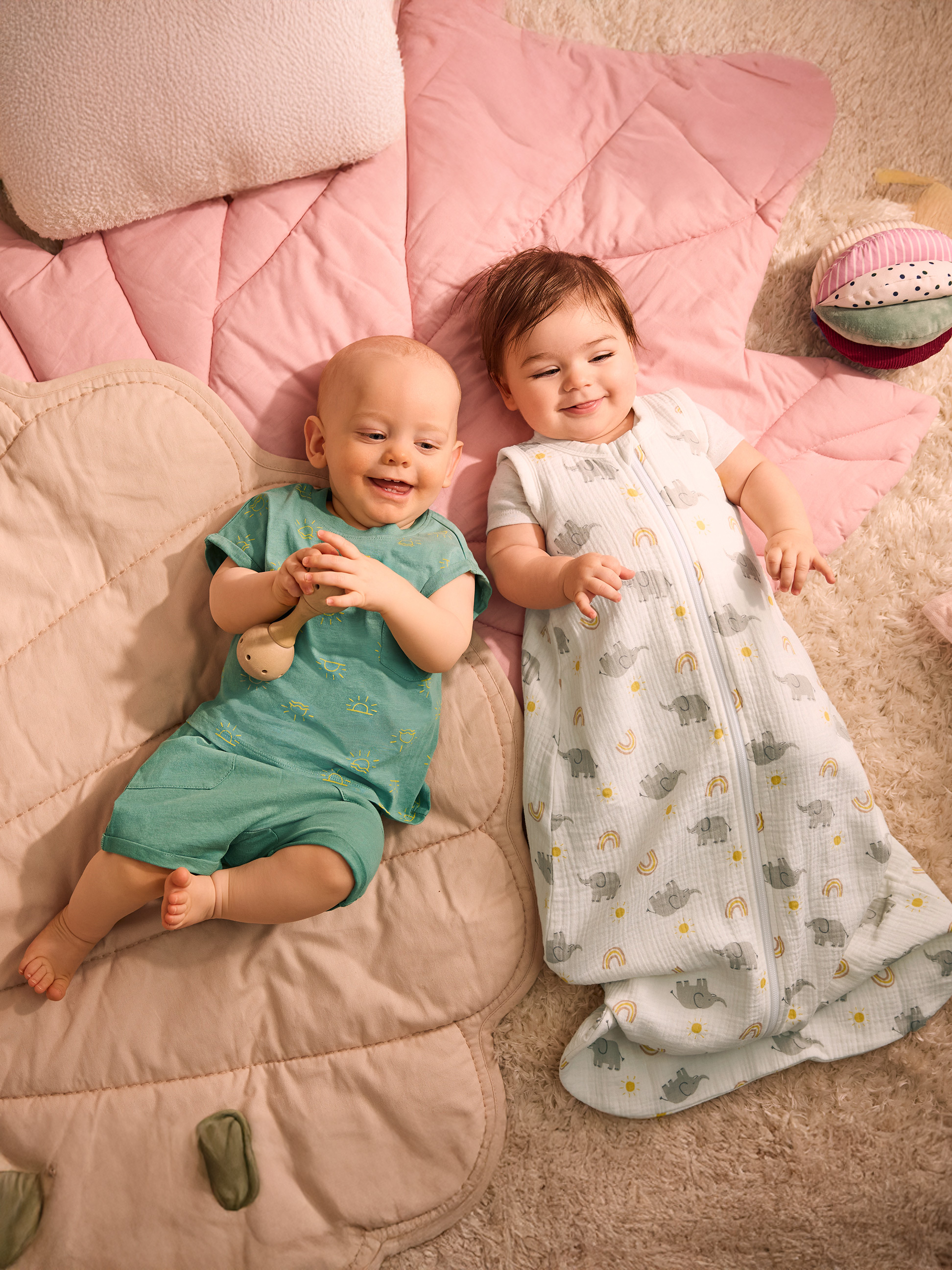 Deux bébés allongés sur une couverture rose, l'un en vêtements verts, l'autre dans une gigoteuse à éléphants.