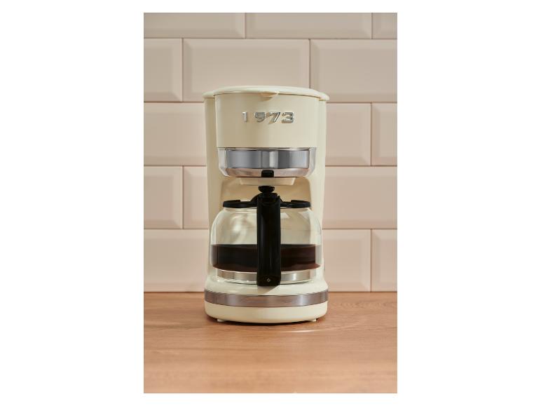 Cafetière crème avec carafe sur un comptoir en bois, devant des carreaux beiges.