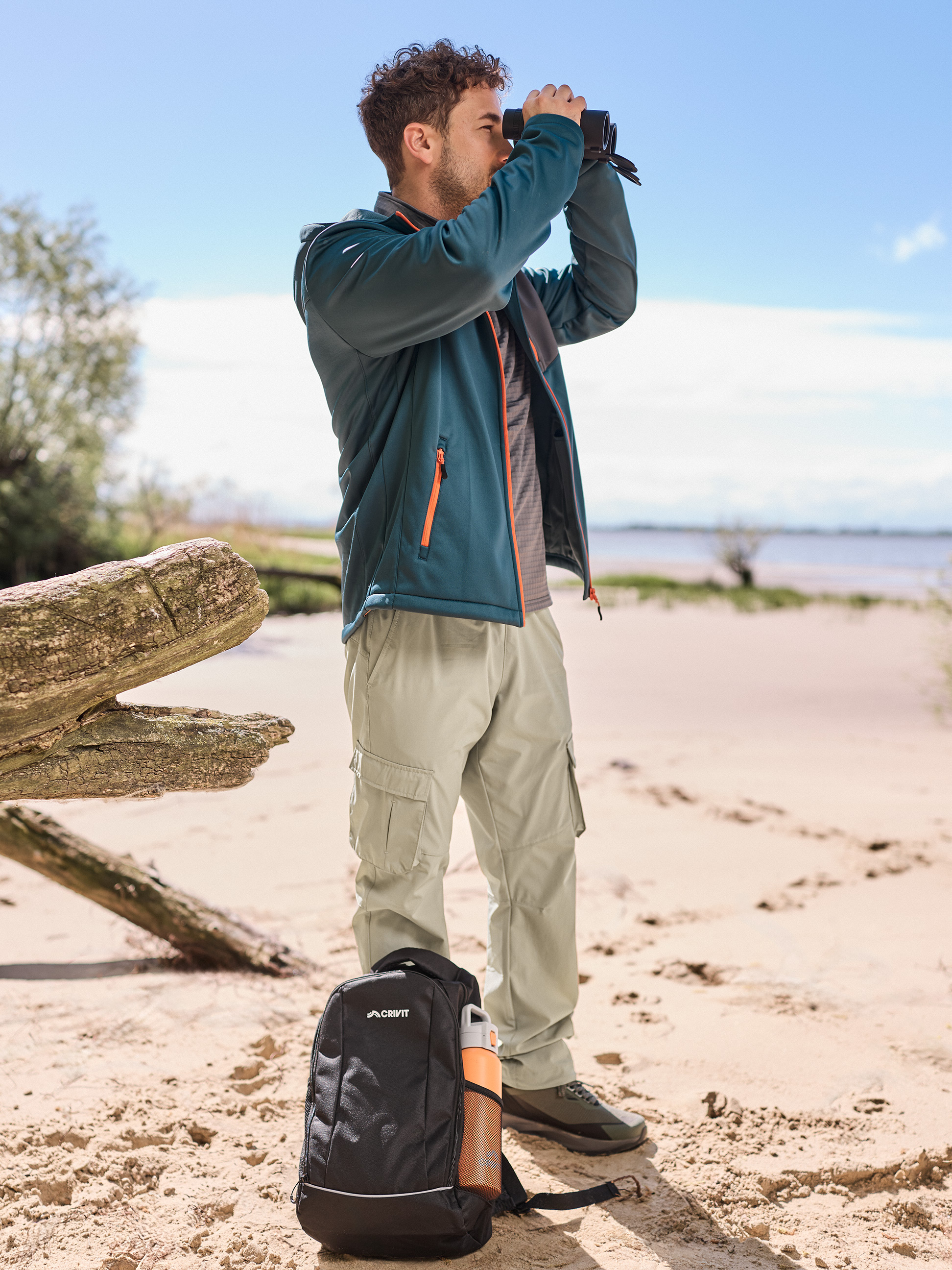 Homme en veste et pantalon de randonnée, jumelles, sac à dos et bouteille d'eau sur la plage