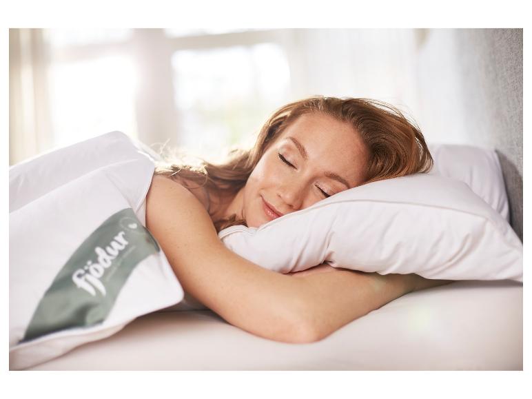 Femme dort sur oreiller et couette blancs avec logo Fjödur dans chambre ensoleillée.