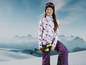Une fille vêtue d'une tenue de ski tient des bâtons de ski devant une chaîne de montagnes enneigées.