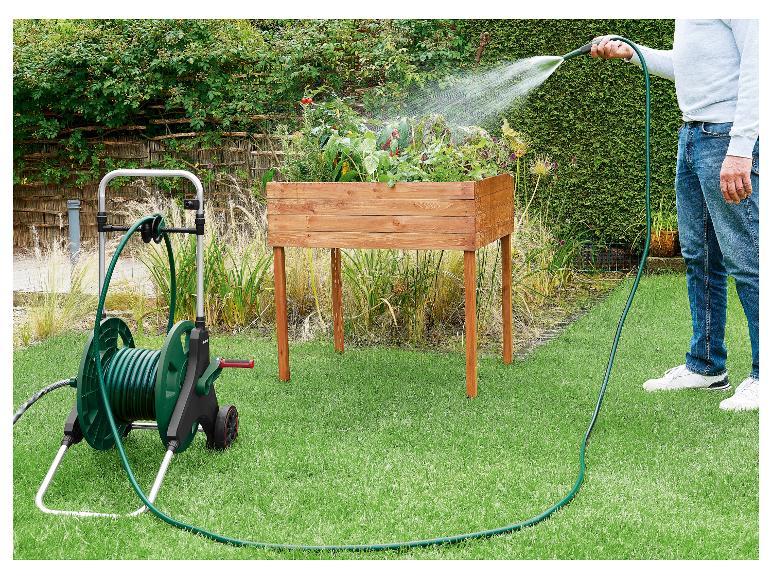 Homme arrosant des plantes dans un bac en bois avec un tuyau d'arrosage vert sur un chariot.