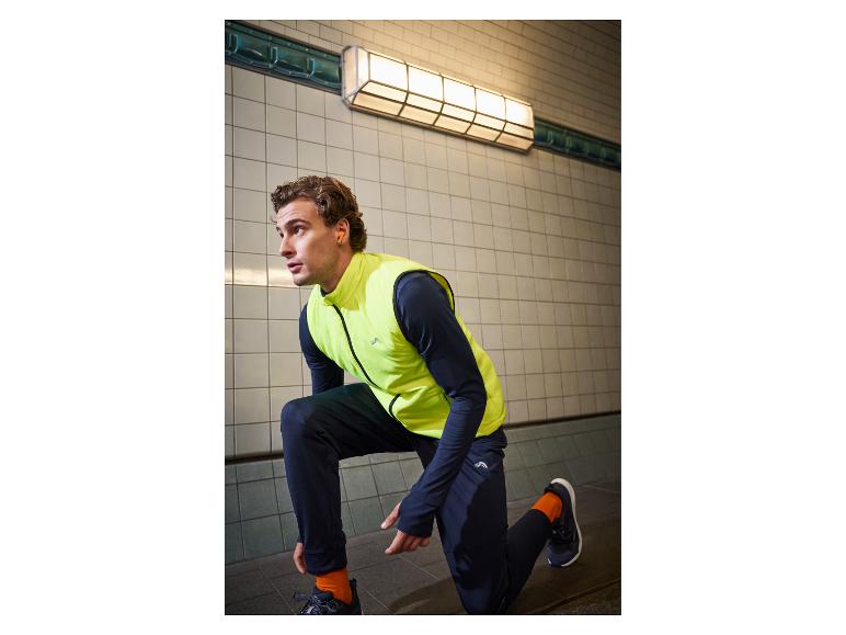 Homme en gilet de sport jaune et pantalon noir, accroupi dans un passage souterrain.