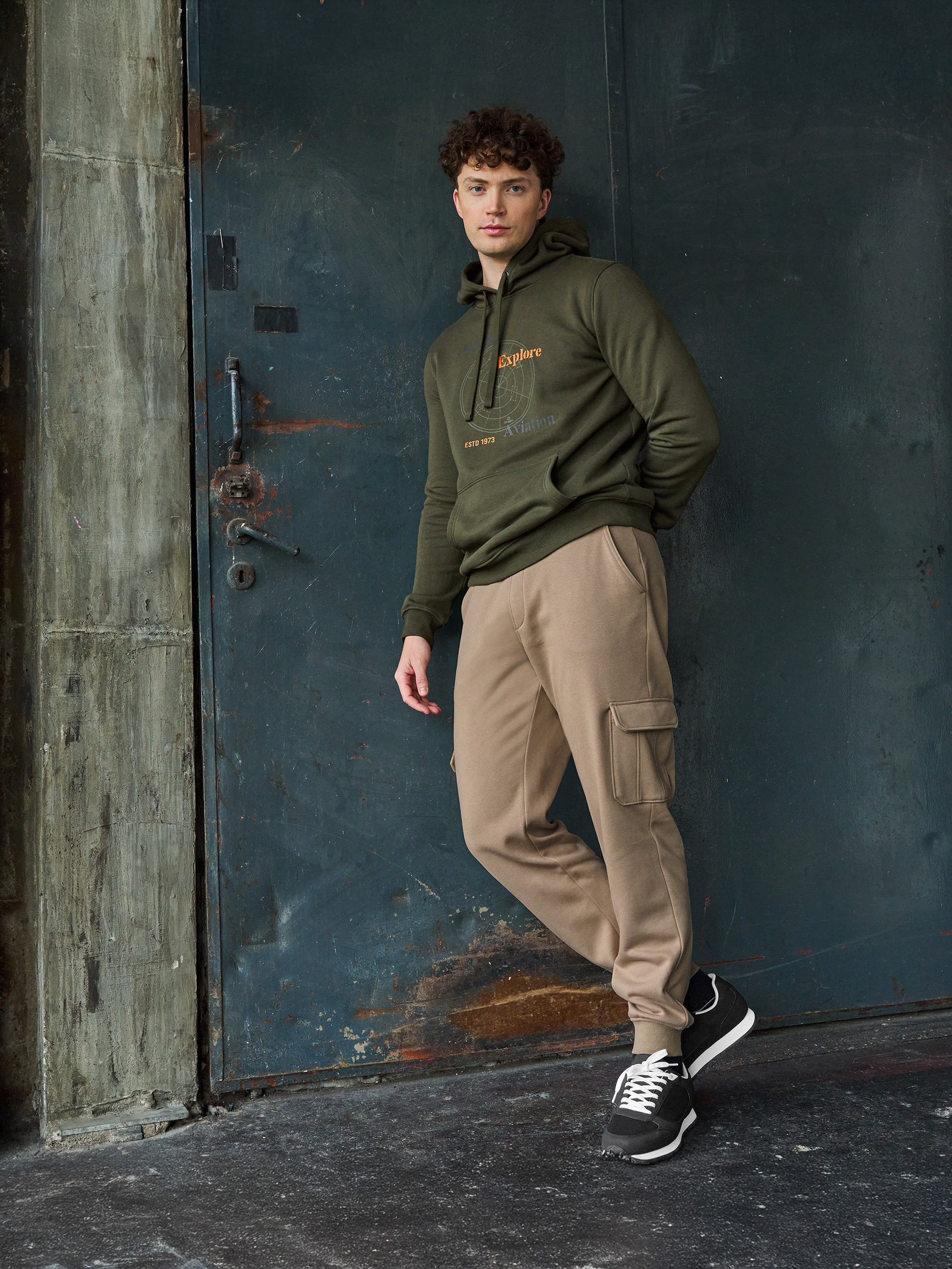 Homme en sweat à capuche vert olive 'Explore Aviation' et pantalon de jogging beige.