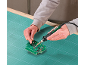 Mains soudant une carte de circuit imprimé avec un fer à souder sur un tapis de découpe vert.