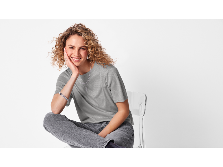 Une femme en t-shirt gris clair et en jeans pose sur une chaise transparente.