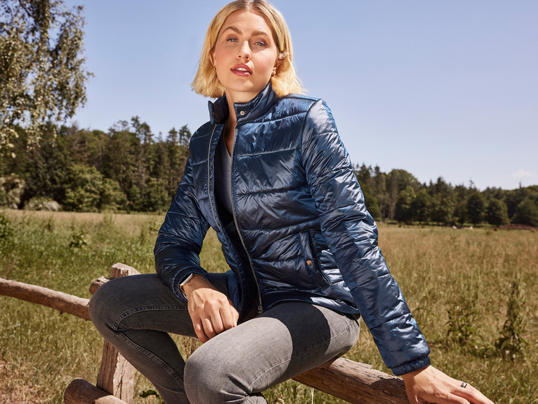 Une femme portant une veste matelassée bleue et un jean est assise sur une clôture en bois.
