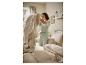 Femme en pyjama vert clair faisant un lit avec une literie beige clair dans une chambre lumineuse.