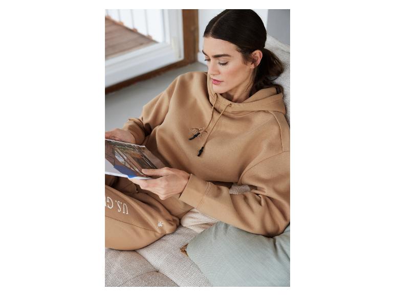 Une femme en sweat-shirt à capuche beige et pantalon de survêtement de U.S. Polo Assn. lit un magazine.