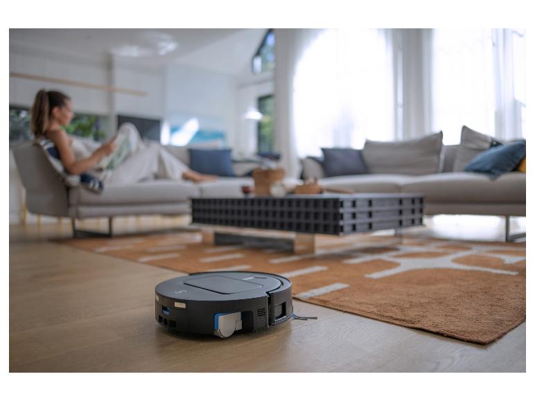 Aspirateur robot sur parquet dans un salon avec une femme lisant en arrière-plan.