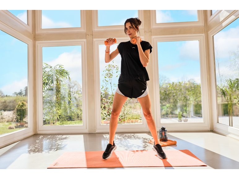 Une femme en tenue de sport s'entraîne avec des haltères sur un tapis orange.