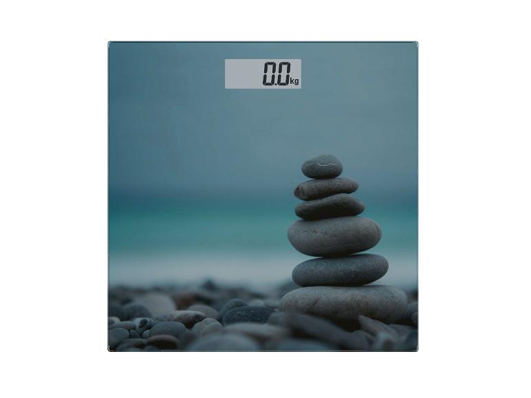 Pèse-personne numérique avec affichage 0.0 kg et pierres empilées sur la plage.