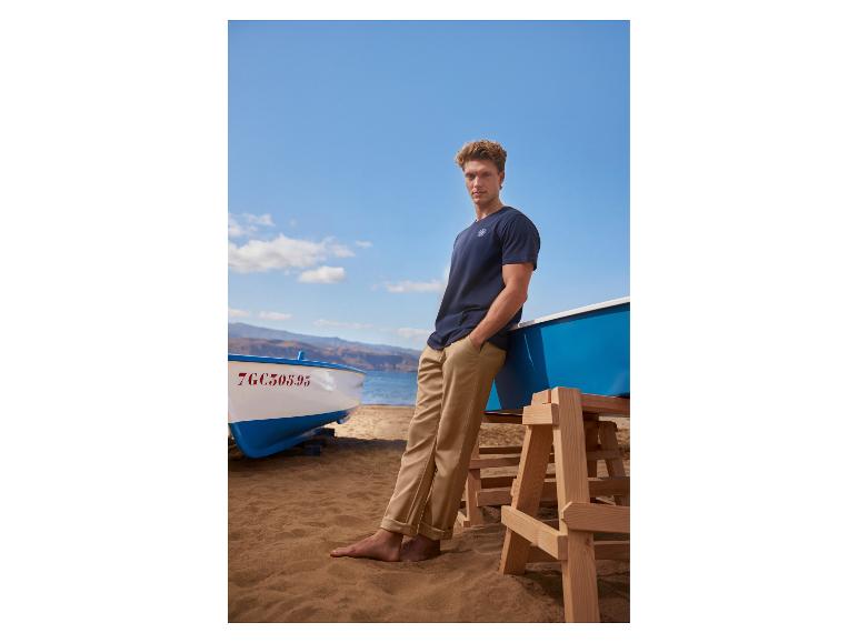 Homme en t-shirt bleu marine et pantalon beige sur la plage, appuyé contre un bateau.