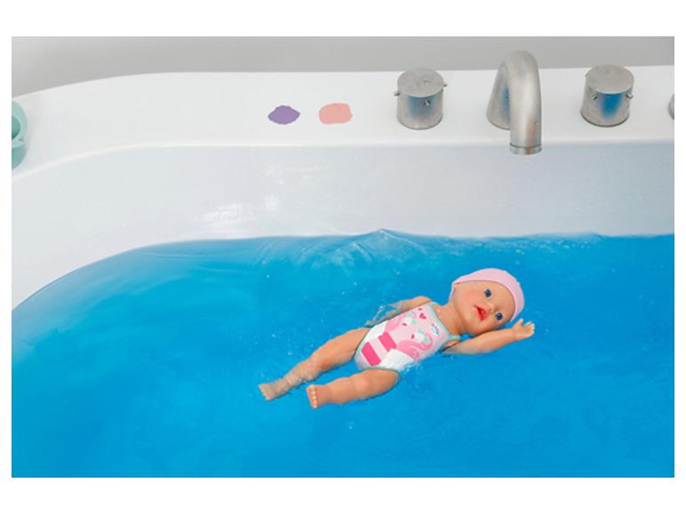 Poupée bébé nageant dans l'eau bleue d'une baignoire, vêtue d'un maillot de bain rose.