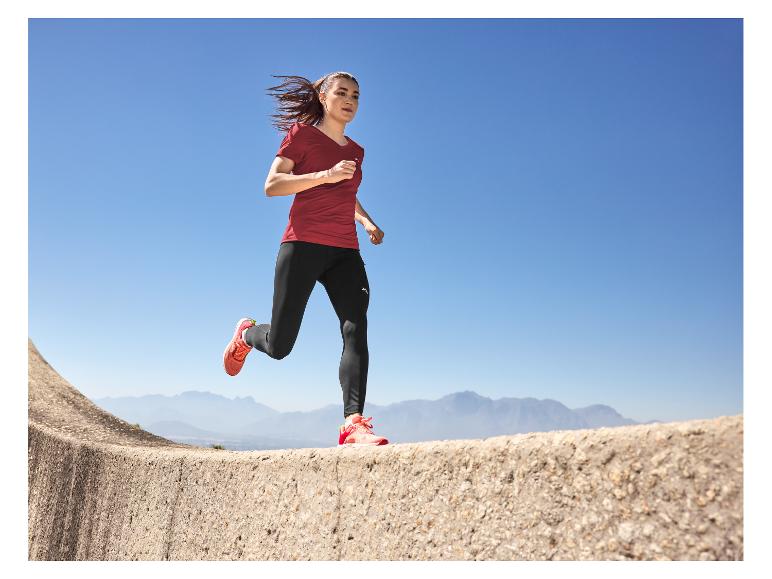 Femme en t-shirt rouge et legging noir courant en extérieur avec des montagnes en arrière-plan.