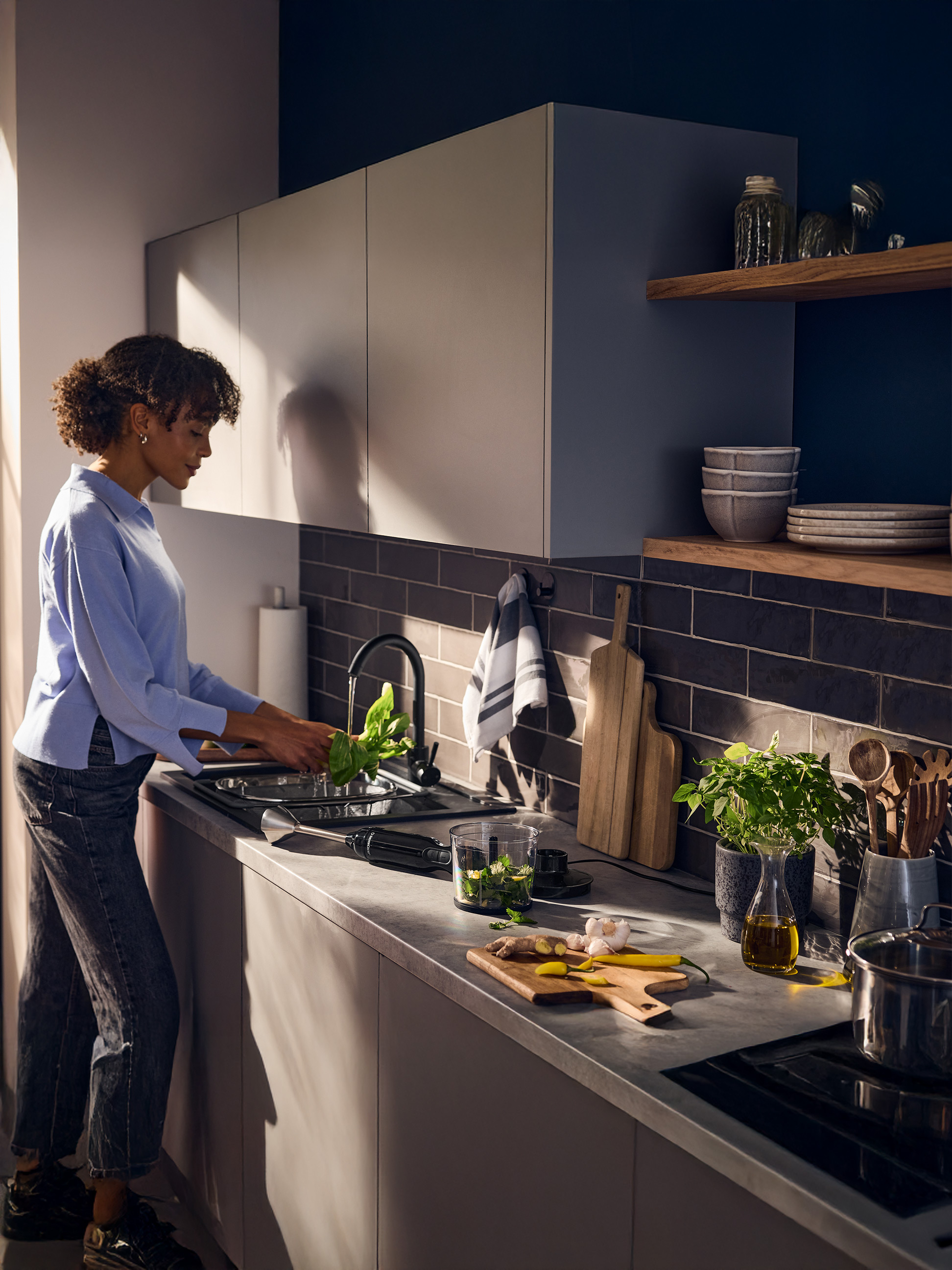 Femme lavant des herbes fraîches dans l'évier de la cuisine, avec mixeur plongeant et ingrédients sur le comptoir.