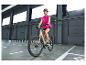 Femme en gilet rose et casque, faisant du vélo électrique en intérieur.