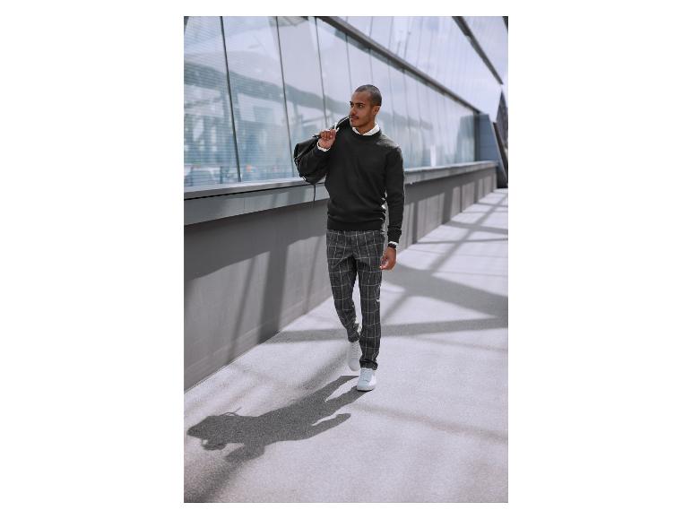 Un homme en pull noir, pantalon à carreaux et baskets blanches marche dans la rue.