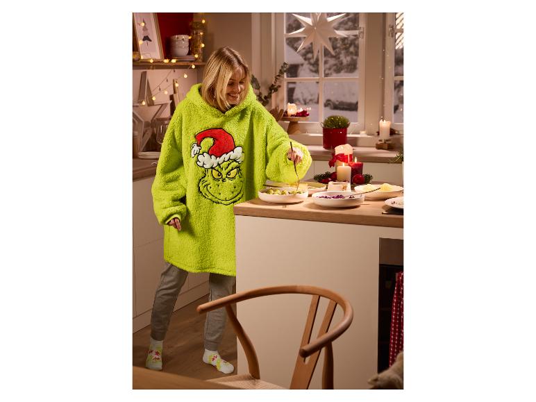 Femme en sweat à capuche oversize sur le thème du Grinch préparant de la nourriture dans une cuisine décorée pour Noël.