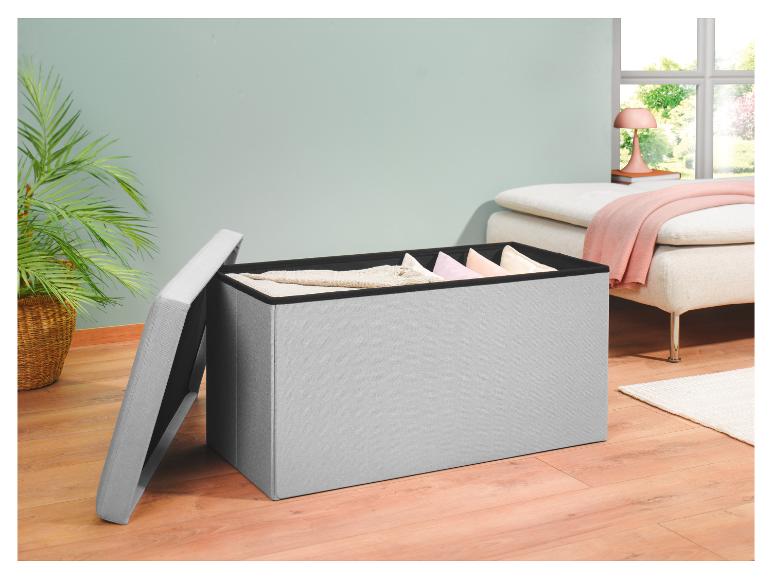 Un pouf de rangement en tissu gris avec un couvercle ouvert, révélant des coussins et des couvertures à l'intérieur, se trouve sur un sol en bois clair.