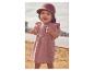 Bébé en robe rose et chapeau de soleil, debout sur une plage de sable.