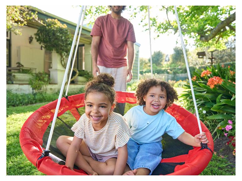 Deux enfants sourient dans une balançoire de jardin rouge, avec papa en arrière-plan.