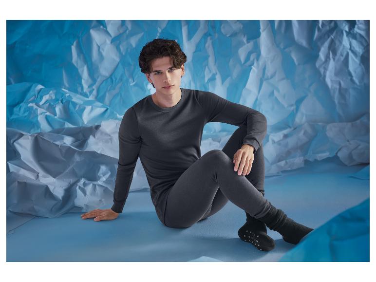 Homme en ensemble de sous-vêtements thermiques gris foncé, assis sur fond bleu.