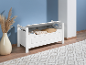 Banc de rangement en bois blanc avec couvercle ouvert, révélant des couvertures et des livres, se trouve sur un sol en bois clair avec un tapis.