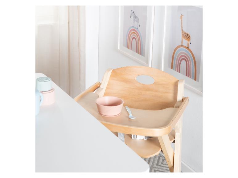 Chaise haute en bois avec un bol rose et une cuillère, à côté d'une table blanche avec de la vaisselle pour bébé.