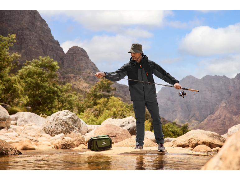 Homme pêchant dans un ruisseau de montagne, portant une veste imperméable, un pantalon et un chapeau, avec un sac de pêche.