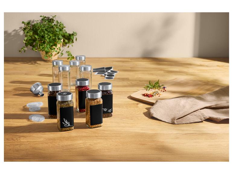 Épices dans des bocaux en verre avec étiquettes noires, entonnoir et herbes fraîches sur table en bois.