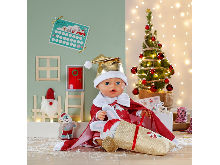 Poupée bébé en costume de Père Noël avec une hotte de cadeaux devant un décor festif.