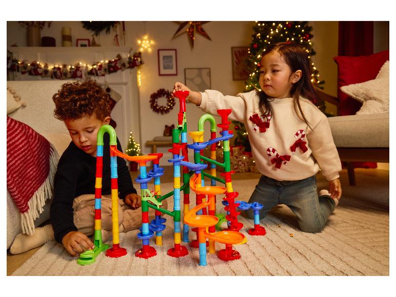 Deux enfants jouent avec un circuit de billes coloré sur un tapis dans une pièce décorée pour Noël.