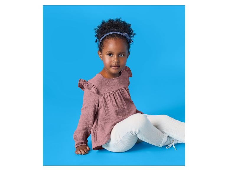 Petite fille aux cheveux bouclés, portant un chemisier rose et un pantalon blanc, assise sur fond bleu.