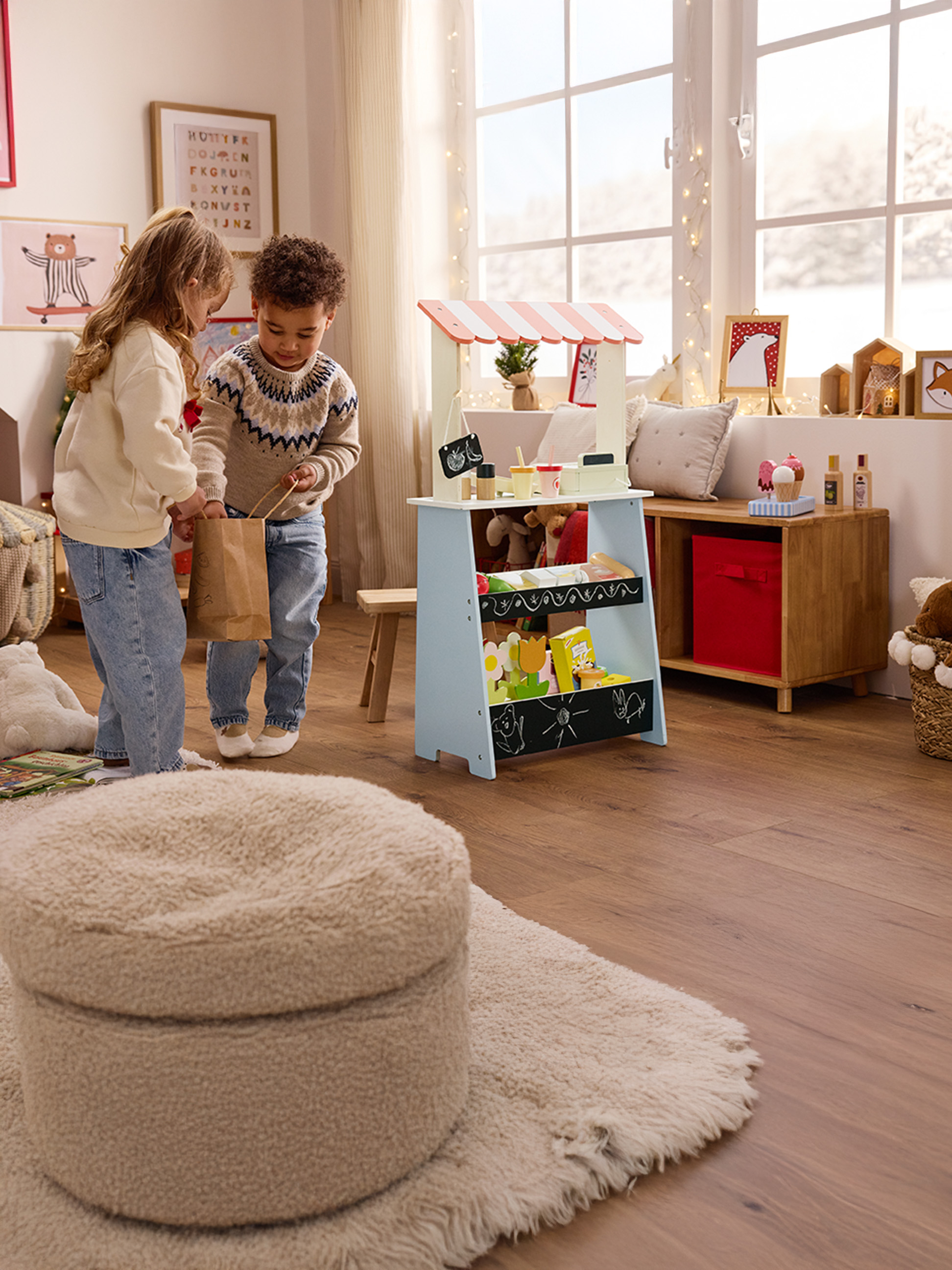 Deux enfants jouent avec un étal de marché en bois, une fille tenant un sac en papier.