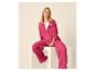 Femme souriante en chemise et pantalon large fuchsia, assise sur un bloc blanc.