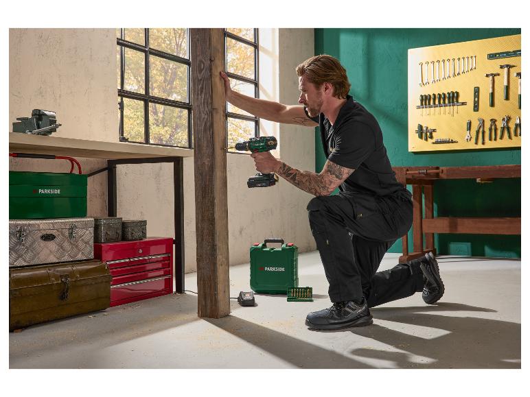 Homme utilisant une perceuse sans fil pour travailler sur une poutre en bois dans un atelier, entouré de divers outils.