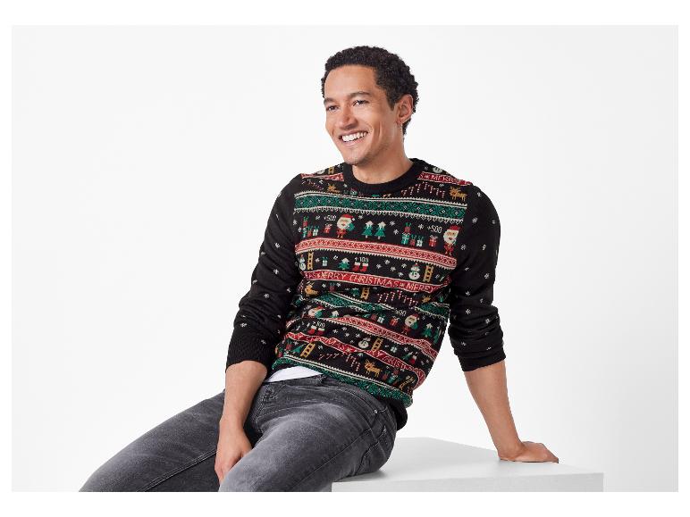 Homme souriant portant un pull de Noël noir avec des motifs festifs et un jean gris.