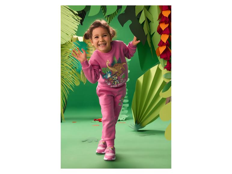 Fille en survêtement rose imprimé dinosaures et baskets, souriante dans un décor de jungle en papier.
